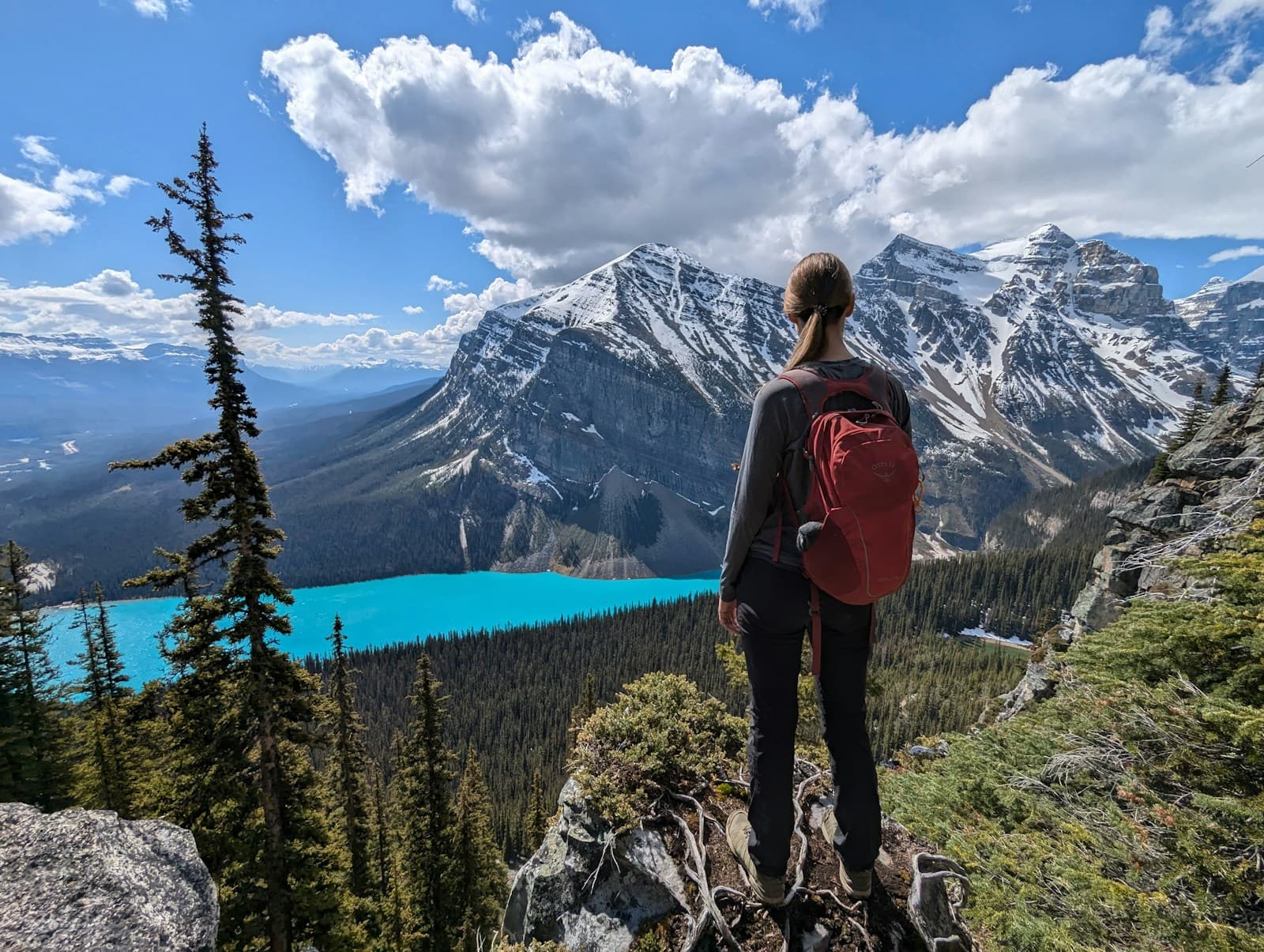 A hiker with a red backpack overlooking turquoise Lake Louise and snow-covered mountains
